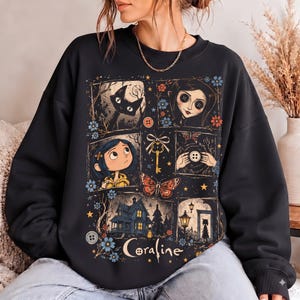 May include: A black crewneck sweatshirt featuring a graphic print with the text Coraline. The design includes panels showing a black cat, a character with button eyes, a key, a butterfly, a house, and blue flowers on a dark background.