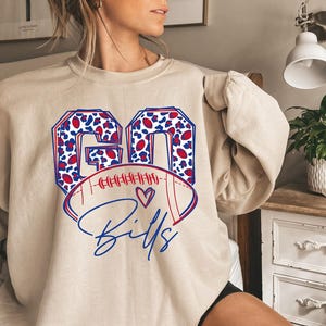 Op de afbeelding: Een beige sweater met ronde hals voorzien van een grafische print met de tekst GO BILLS in blauw en rood. De letters GO zijn gevuld met een blauw en rood luipaardpatroon. Een illustratie van een football met een klein hartje staat onder de tekst.