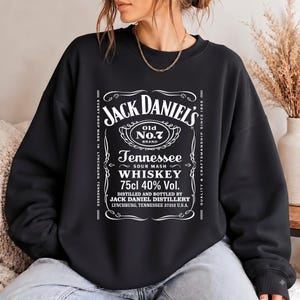 May include: A black crewneck sweatshirt featuring a white graphic print of the Jack Daniel's Old No. 7 Tennessee Sour Mash Whiskey label, including text details like 75cl 40% Vol. and the distillery location in Lynchburg, Tennessee.