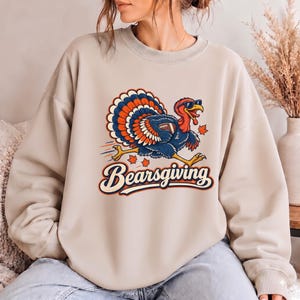 May include: A beige crewneck sweatshirt featuring a graphic of a turkey wearing sunglasses and carrying a football. The turkey is surrounded by falling maple leaves, and the text "Bearsgiving" is printed below in a bold, retro-style font.