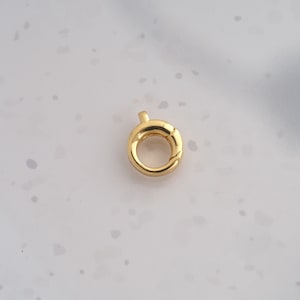May include: A small, circular gold-toned spring ring clasp used for jewelry making. The component features a smooth, polished metallic finish and a functional spring-loaded mechanism with a tiny tab for opening the closure loop.
