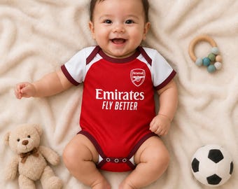May include: A red and white baby bodysuit featuring the Arsenal football club crest and the text Emirates FLY BETTER in white. The garment has short sleeves, a round neckline, and snap closures at the bottom for easy diaper changes.