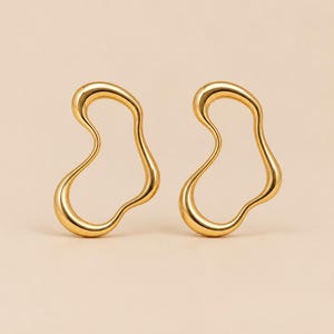 May include: A pair of gold-toned earrings featuring an irregular, fluid, and organic open-loop shape. The metal surface is smooth and polished, reflecting light along the curved, continuous edges of the abstract, hollow, and rounded design.