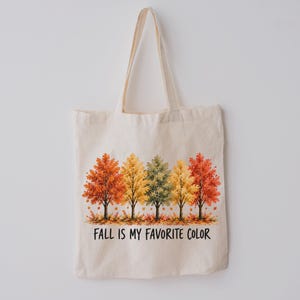 Op de afbeelding: Een beige canvas draagtas met een grafische print van vijf bomen met herfstkleurige bladeren in tinten oranje, geel en groen. Onder de bomen staat de tekst FALL IS MY FAVORITE COLOR in een eenvoudig zwart schreefloos lettertype.
