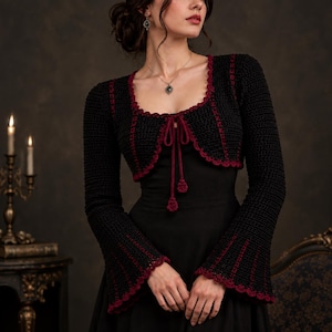 May include: A black crochet bolero jacket featuring long flared sleeves and a deep red trim along the edges. The garment has a square neckline and a front tie closure with two dangling red tassels. The crochet pattern is consistent throughout.