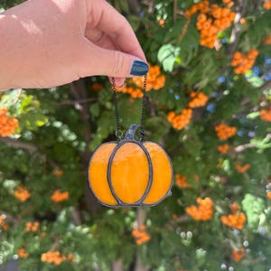 Könnte beinhalten: Ein Kürbis-Ornament aus Buntglas mit leuchtend orangefarbenen Glassegmenten, die durch dunkle Metalllötstellen verbunden sind. Der Kürbis hat einen kleinen dunklen Stiel an der Oberseite und ist an einer dünnen schwarzen Metallkette aufgehängt.