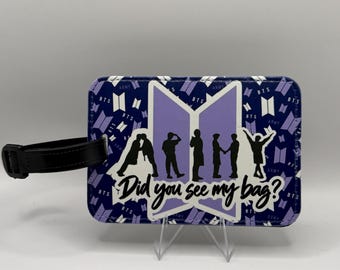 May include: A rectangular luggage tag with a purple and white background featuring the BTS logo and the text 'Did you see my bag?'. The centre displays black silhouettes of seven figures against a light purple BTS logo, with a black strap attached.