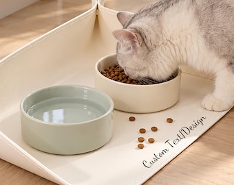 Può includere: Un tappetino per l'alimentazione di animali domestici color crema, pieghevole con angoli a bottone, dotato di due ciotole rotonde per cibo e acqua. Il tappetino riporta la scritta Custom Text/Design in nero. Un gatto grigio e bianco sta mangiando.