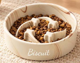 May include: A round, beige and brown marble-patterned ceramic slow feeder bowl for pets. The bowl features an internal star-shaped obstacle design and the word Biscuit printed in white script on the front exterior, filled with brown dry kibble.