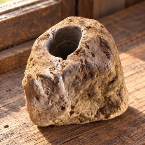 May include: A natural tan-colored stone vessel with a rough, porous, and uneven exterior texture. The object features a single, smooth, cylindrical opening carved into the top center, revealing a darker gray interior surface within the rock.