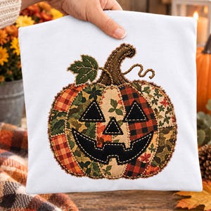 May include: A white t-shirt featuring a large, central pumpkin applique. The pumpkin is constructed from various fabric patches in orange, brown, and green, including plaid and floral patterns. It has a black fabric jack-o'-lantern face.