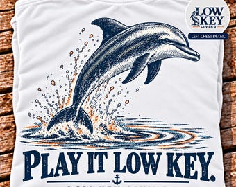 May include: A white t-shirt featuring a dark blue graphic of a dolphin leaping from water with orange splashes. Below the dolphin, the text reads "PLAY IT LOW KEY." and "LOW KEY LIVING" with an anchor icon. A circular inset shows a pelican logo.