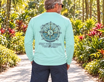 May include: A light mint green long-sleeve shirt featuring a large graphic on the back. The design displays a detailed nautical compass with a pelican in the center, surrounded by the text STAY YOUR COURSE and LOW KEY LIVING.