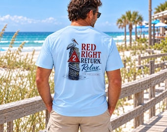 May include: A light blue short-sleeve t-shirt featuring a graphic on the back of a pelican perched on a wooden piling with a red triangular sign marked with the number 2. The text reads RED RIGHT RETURN. Relax. LOW PET LIVING.