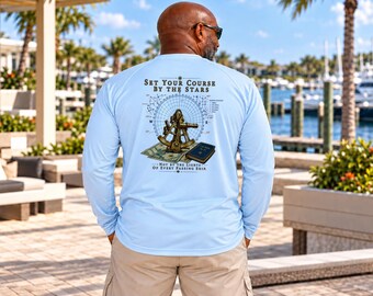 May include: A light blue long-sleeve shirt featuring a graphic on the back with a sextant, a map, and a book. The text reads: Set Your Course By The Stars, Not By The Lights Of Every Passing Ship. The design includes a compass rose.