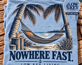 May include: A light blue t-shirt featuring a graphic of a hammock strung between two palm trees on a beach. The design includes a drink on a wooden post, the text "NOWHERE FAST." and "LOW KEY LIVING," and a small anchor icon.