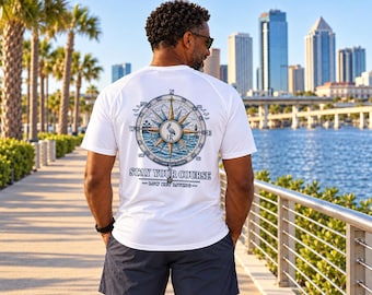 May include: A white short-sleeved t-shirt featuring a large graphic on the back of a nautical compass with a pelican in the center. The text reads STAY YOUR COURSE and LOW KEY LIVING. The design includes blue and gold accents with waves.
