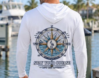 May include: A white long-sleeve hooded shirt featuring a large graphic on the back of a nautical compass rose with a pelican in the center. The text reads "STAY YOUR COURSE" and "LOW KEY LIVING" with "DON'T FIGHT EVERY CURRENT" below the compass.