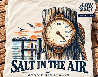 May include: An ivory t-shirt featuring a graphic of a wooden piling with a gauge labeled Dry, Nice, Humid, Sticky, and Swampy. The scene includes a sunset, a pier, birds, and rope. Text reads: Salt in the air. Good vibes always. Low Key Living.