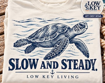 May include: An ivory t-shirt featuring a dark blue graphic of a sea turtle swimming in waves. Below the turtle, the text reads "SLOW AND STEADY." and "LOW KEY LIVING" with a small anchor icon. A circular inset shows a pelican logo and "LOW KEY LIVING".