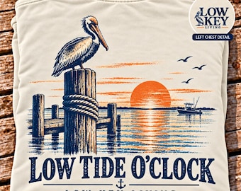 May include: An ivory t-shirt featuring a graphic of a pelican perched on a wooden piling at sunset. The design includes a boat, birds, and the text LOW TIDE O'CLOCK and LOW KEY LIVING. A circular inset shows the LOW KEY LIVING logo.