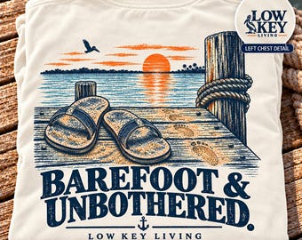 May include: An ivory t-shirt featuring a graphic of a wooden pier at sunset with a pair of sandals, footprints, a piling with rope, and a flying pelican. The text reads BAREFOOT & UNBOTHERED. LOW KEY LIVING with an anchor icon.