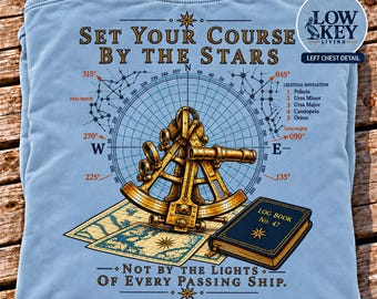 May include: A light blue t-shirt featuring a graphic of a gold sextant, a blue log book, and a celestial navigation map. The text reads: SET YOUR COURSE BY THE STARS, NOT BY THE LIGHTS OF EVERY PASSING SHIP, and includes a list of constellations.