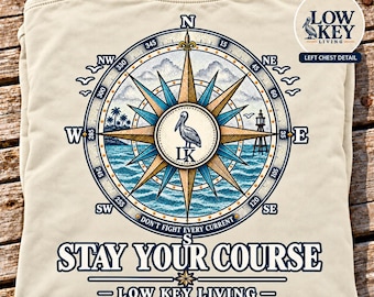 May include: An ivory t-shirt featuring a large back graphic of a compass rose with a pelican in the center, ocean waves, palm trees, and a lighthouse. The text reads "DON'T FIGHT EVERY CURRENT", "STAY YOUR COURSE", and "LOW KEY LIVING".