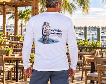 May include: A white long-sleeve shirt featuring a graphic on the back of a wooden piling in water with a sign that reads IDLE SPEED and the text No Wake. No Hurry. LOW KEY LIVING. The shirt is worn with dark gray shorts in an outdoor setting.