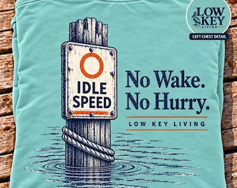 May include: A light teal t-shirt featuring a graphic of a wooden piling in water with a rope wrapped around it. A sign on the piling reads IDLE SPEED with an orange circle. Text reads No Wake. No Hurry. LOW KEY LIVING and a logo.