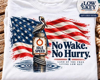 May include: A white t-shirt featuring a graphic of a pelican perched on an idle speed sign, set against a waving American flag. The text reads No Wake. No Hurry. Land of the Free Living Low Key. A circular inset shows the Low Key Living logo.