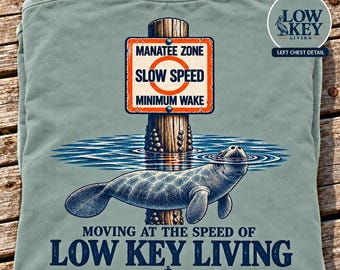 May include: A sage green t-shirt featuring a graphic of a manatee swimming near a wooden post with a sign that reads MANATEE ZONE SLOW SPEED MINIMUM WAKE. Below the manatee, the text reads MOVING AT THE SPEED OF LOW KEY LIVING.