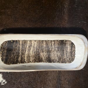 Peut inclure: Un bol à pâte en bois de couleur claire, de forme allongée, doté d'un insert rectangulaire en peau de vache marron et blanche. La peau est fixée à la base intérieure du bol par une série de petits clous métalliques régulièrement espacés.