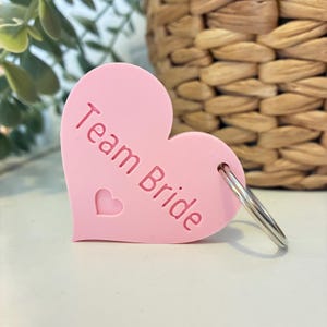 以下が含まれることがあります： Team Brideというエンボス加工の文字と小さなハートの切り抜きが施された、ライトピンクのハート型キーホルダー。キーホルダーにはシルバーカラーの金属製二重リングが付いており、鍵をまとめたりバッグに取り付けたりするのに機能的です。
