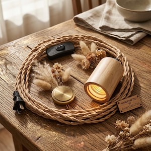 May include: A light-colored wooden cylindrical lamp socket attached to a braided beige cord with a black power switch and plug. The cord is coiled and decorated with dried flowers and a small brass disc. A wooden tag reads Handcrafted by AvenzaGifts.