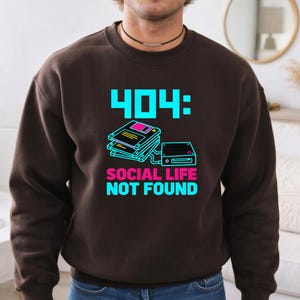 May include: A dark brown crewneck sweatshirt featuring a graphic print with the text 404: SOCIAL LIFE NOT FOUND in bright cyan and pink. The design includes a stack of three floppy disks and a disk drive in a retro computer style.