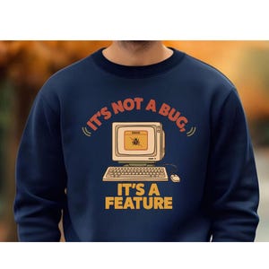 May include: A navy blue crewneck sweatshirt featuring a graphic of a vintage computer monitor and keyboard. The screen displays an error box with a bug icon. The text reads, "IT'S NOT A BUG, IT'S A FEATURE" in orange and yellow lettering.