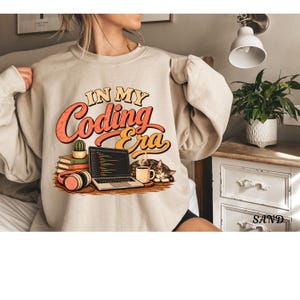 May include: A sand-colored crewneck sweatshirt featuring a graphic print with the text "IN MY Coding Era" in orange and yellow. The design includes a laptop displaying code, a stack of books, a cactus, headphones, a mug, and a sleeping cat.