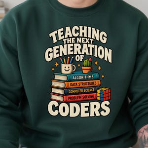 May include: A dark green sweatshirt featuring a graphic print with the text "TEACHING THE NEXT GENERATION OF CODERS". The design includes a stack of four books labeled "ALGORITHMS", "DATA STRUCTURES", "COMPUTER SCIENCE", and "PROBLEM SOLVING", a mug with a smiley face, a small cactus, and a colorful cube.