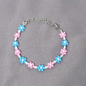 Puede incluir: Una pulsera de cuentas con flores alternas en azul claro y rosa claro, cada una con una cuenta blanca en el centro. La pulsera está unida por una cadena de metal plateado con un cierre de mosquetón para un ajuste de tamaño regulable.