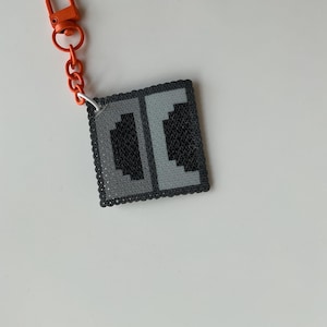 May include: A square pixel art keychain featuring the letters DC in a dark gray and light gray color scheme. The keychain is attached to a bright orange plastic lobster clasp and chain, creating a distinct contrast with the square pendant.