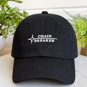 Peut inclure: Une casquette de baseball noire avec le texte brodé en blanc CHAIN BREAKER, accompagné d'un graphique en forme d'étoile à quatre branches à gauche des mots. La casquette présente une calotte structurée, une visière courbée et un bouton au sommet.
