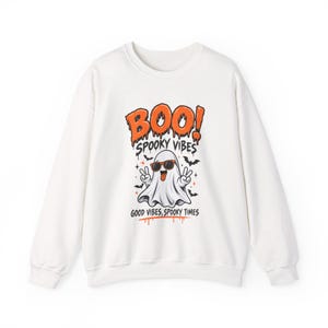 Puede incluir: Una sudadera blanca de manga larga con un estampado gráfico de un fantasma con gafas de sol naranjas haciendo el signo de la paz. El texto dice BOO! SPOOKY VIBES y GOOD VIBES, SPOOKY TIMES en naranja y negro, rodeado de pequeños murciélagos y estrellas.