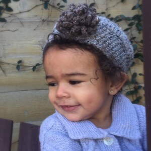 Puede incluir: Un niño pequeño con un cárdigan de punto de color lila claro con cierre de botón blanco. El niño lleva un gorro de punto gris y marrón con una flor en la parte superior.