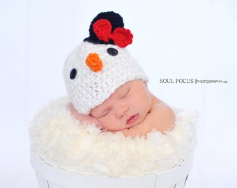 Puede incluir: Un bebé recién nacido con un gorro de punto blanco de muñeco de nieve con un pompón rojo y negro en la parte superior. El bebé está durmiendo en una cesta blanca y esponjosa.