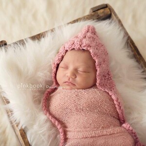 May include: A sleeping baby wearing a pink crocheted hat and swaddled in a pink blanket. The baby is lying in a wooden basket lined with white fur.