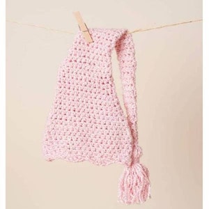 Op de afbeelding: Een lichtroze, gehaakte baby muts met een lange, puntige staart en kwast. De muts hangt aan een waslijn met een houten knijper. De textuur van de muts is zichtbaar en de kleur is zacht en pastel.