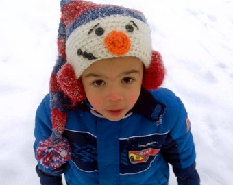 Puede incluir: Un niño con un abrigo de invierno azul y un gorro de crochet blanco con una cara de muñeco de nieve. El gorro tiene detalles en rojo, blanco y azul. El niño está de pie en la nieve.