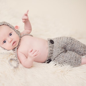 May include: A baby wearing a gray crocheted hat with a pink flower, a gray crocheted outfit, and lying on a white fluffy surface.