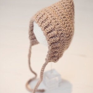 Peut inclure: Un bonnet de bébé en crochet marron avec de longues attaches. Le bonnet est présenté sur un support blanc.
